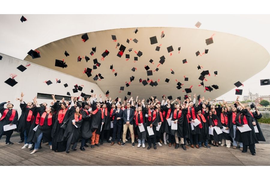 Photo de groupe. Les 160 diplômés, en tenue d'apparat (toge noire, écharpe rouge), lancent leur chapeau en l'air devant la "soucoupe volante", le bâtiment emblématique en forme de coupole renversée d'UniLaSalle Amiens